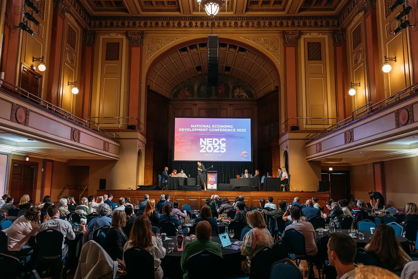 View across delegates seated at round tables looking towards speakers presenting on stage in front of an Economic Development Australia banner at Newcastle City Hall.