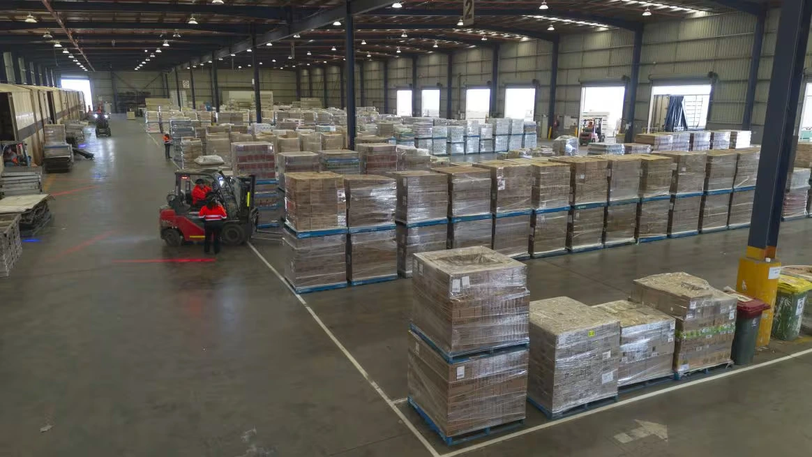 A large warehouse with tall shelves, stacked pallets and workers driving forklifts through wide, organised aisles and loading bays.