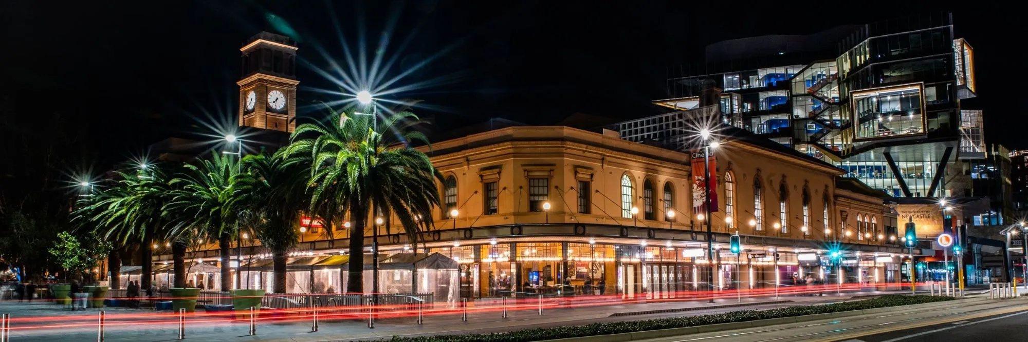 Civic Theatre Newcastle at night with illuminated clock tower, palm trees, and modern buildings in the background.