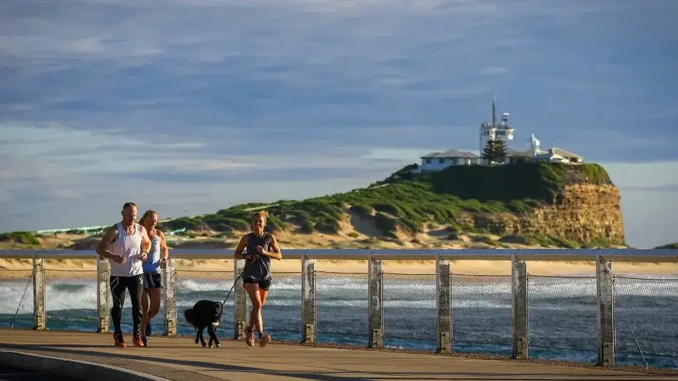 A group of three people and their dog enjoy running along Nobbys Beach, with the Lighthouse in the background. 