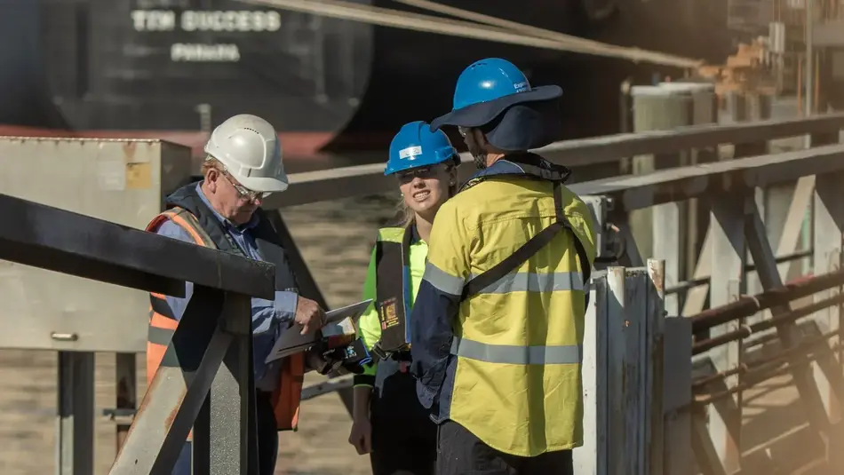 Three workers in safety gear reviewing documents on a wharf at Port of Newcastle with cargo ship in background.
