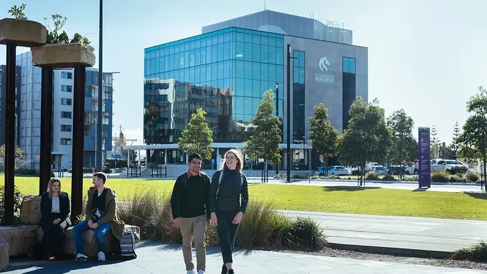 People walking and sitting in front of the University of Newcastle city campus building with glass façade and landscaped area.