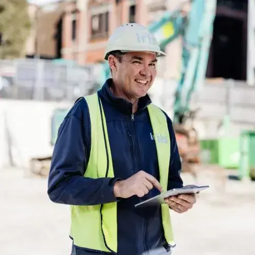 Smiling construction worker wearing a hard hat and safety vest uses a tablet at a construction site.