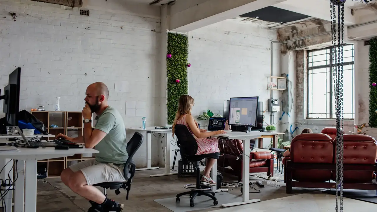 People working at desks in a modern shared office space with exposed brick walls, greenery panels, and large windows.