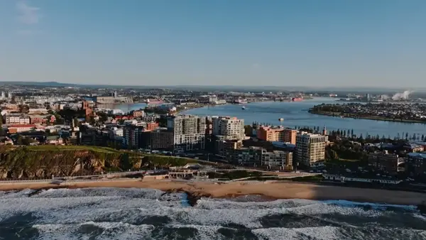Aerial view of Newcastle coastline with ocean waves, city buildings, and harbour in the background under clear skies.