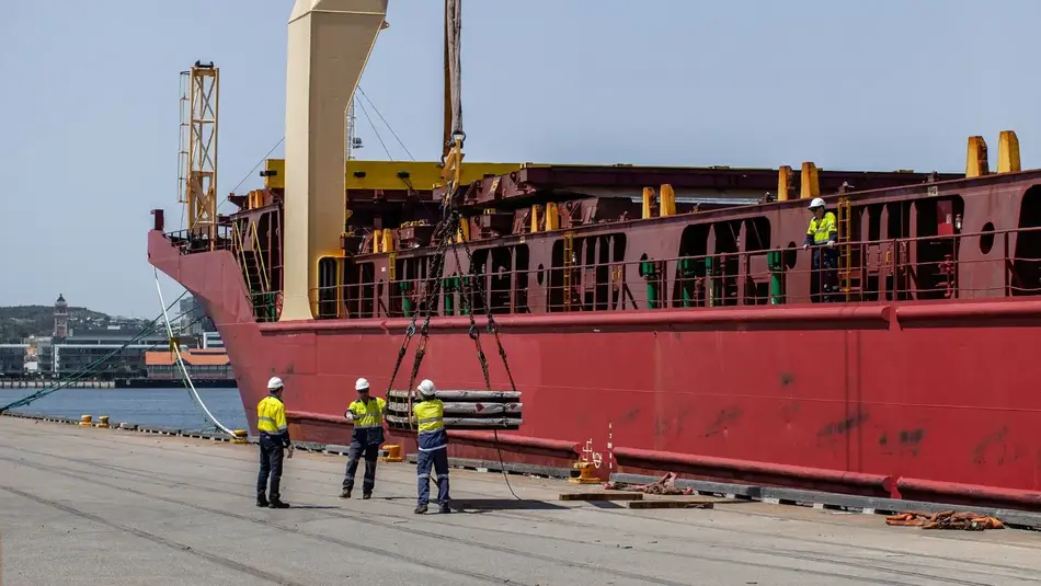 Workers overseeing cargo loading at Port of Newcastle with a large red vessel docked at the wharf.